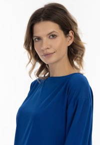 Kék, laza szabású felső széles nyakkivágással és rövid ujjakkal. Sima textúrájú, egyszínű, látható minták vagy díszítések nélkül.