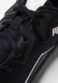 Černá sportovní bota s texturovaným textilním svrškem, černými tkaničkami, plastovými akcenty a bílým zdobením podél podrážky. Obsahuje logo na jazyku.