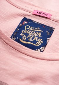 Růžová bavlněná košile s modrou etiketou se zlatým raženým textem "Super Dry" a květinovými akcenty, umístěná poblíž límce.