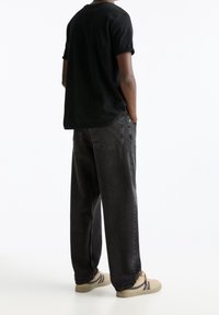 T-shirt nera in cotone con maniche arrotolate, abbinata a jeans grigi scuri a vestibilità ampia. Scarpe beige con accenti scuri completano l'outfit.