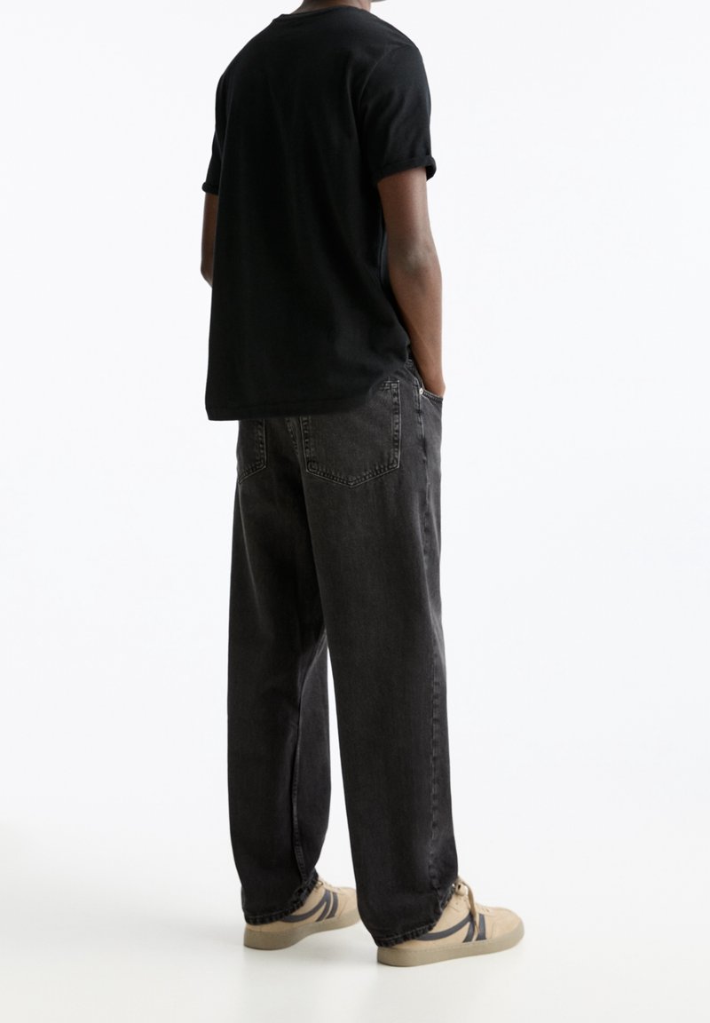 T-shirt nera in cotone con maniche arrotolate, abbinata a jeans grigi scuri a vestibilità ampia. Scarpe beige con accenti scuri completano l'outfit.