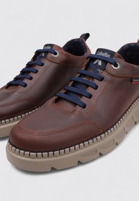 Zapatos de cuero marrón con cordones azul marino, con una suela de goma beige texturizada y costuras decorativas a lo largo del borde. Diseño de puntera redonda.