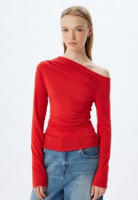 Maglietta rossa a maniche lunghe con design a spalla unica, texture arricciata e silhouette fitted. Abbinata a jeans in denim blu.
