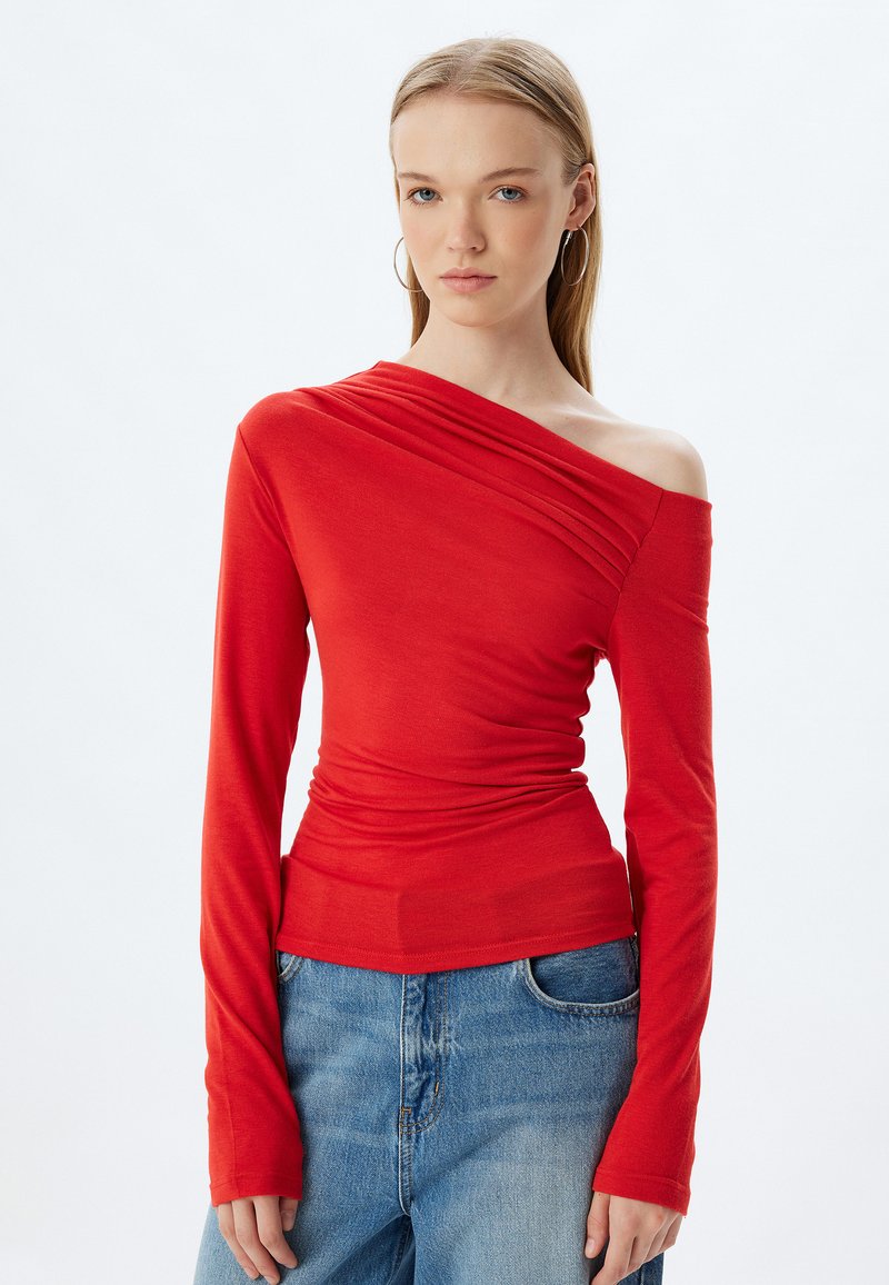 Maglietta rossa a maniche lunghe con design a spalla unica, texture arricciata e silhouette fitted. Abbinata a jeans in denim blu.