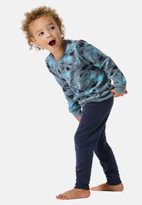 Blaues Langarmhemd mit Dinosauriermotiven, kombiniert mit navy-blauen, weichen Sweatpants. Das Kind steht barfuß und posiert mit den Händen in den Hüften.