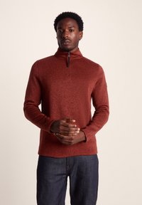 Rust-colored pullover sweater with a high collar, zip neckline, and long sleeves, featuring a textured knit design. Worn with dark blue jeans.
