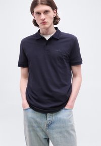 Νεαρός άνδρας με μακριά μαλλιά μέχρι τους ώμους, φορούσε μπλε σκούρο πόλο BOSS και ανοιχτό μπλε τζιν, στέκεται με τα χέρια στις τσέπες μπροστά σε λευκό φόντο.