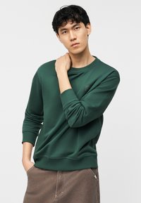 Νεαρός με σκούρα μαλλιά, φορώντας πράσινο φούτερ και καφέ παντελόνι, στέκεται με το ένα χέρι στην τσέπη και το άλλο στον αυχένα.