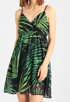 Woman wearing a sleeveless black dress with green tropical leaf print and thin shoulder straps, standing against a plain background.