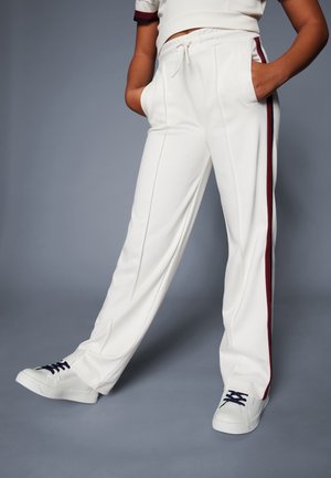 Pantalones deportivos blancos con un corte holgado, que cuentan con bolsillos laterales profundos y una franja burdeos a lo largo de los lados, combinados con zapatillas blancas.