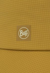 Gorra tipo buff en color amarillo mostaza con un patrón de cuadrícula texturizado. Presenta una etiqueta de logo cuadrada en un tono más claro, centrada en la tela.