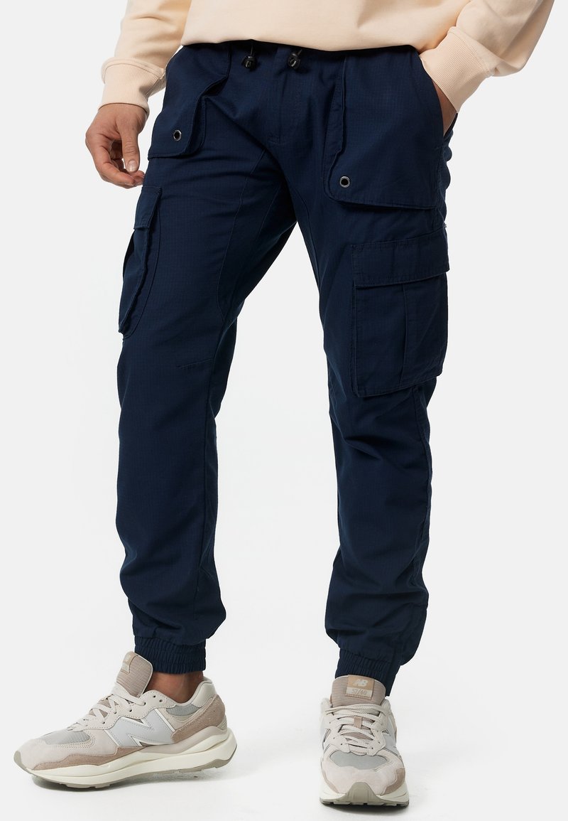 Homme portant un pantalon cargo bleu marine avec plusieurs poches, des chevilles élastiquées, associé à des baskets beiges et blanches.