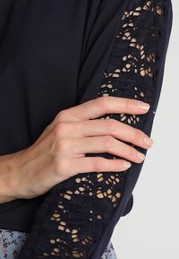 Långärmad marinblå topp med en texturerad spetspanel på ärmen. En hand som vilar på ärmen visar en slät tygkontrast och detalj.