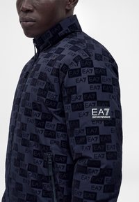 Άνδρας που φοράει ένα ναυτικό μπλε μπουφάν EA7 Emporio Armani με επαναλαμβανόμενο λογότυπο και τσέπη με φερμουάρ στο εμπρός μέρος.