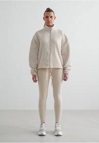 Béžová zipová mikina s oversized střihem a elastickými manžetami. Béžové legíny s hladkou texturou, doplněné o bílé sportovní tenisky s akcenty.