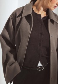 Pārmērīgs pelēks mētelis ar apkakles dekoltē, pārlikts pāri tumšam pogu džemperim. Tam ir gluda tekstūra un melna josta ar sudraba sprādzi.