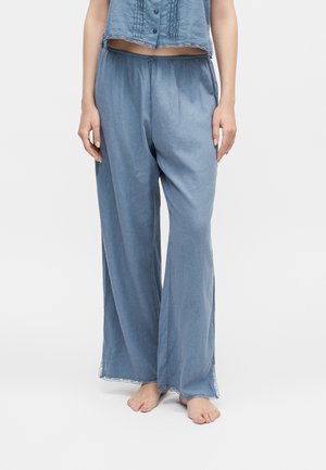 Žena nosiaca voľné, svetlomodré, široké nohavice s vysokým pásom a krátky zladený top, stojaca bosá na svetlom pozadí.