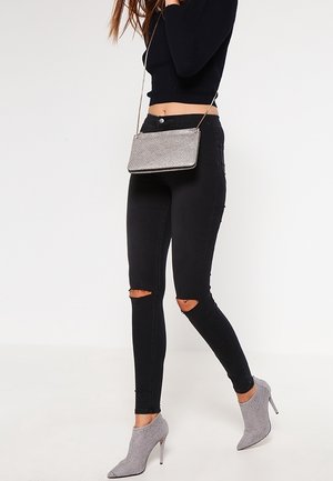 Femme portant un jean skinny noir déchiré, un pull court noir, des bottines grises à talons hauts et un sac bandoulière argenté.