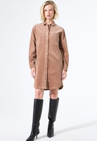 Vestido de camisa de pana marrón con mangas largas, cierre de botones en la parte delantera, dos bolsillos en el pecho y un dobladillo curvado, combinado con botas negras hasta la rodilla.