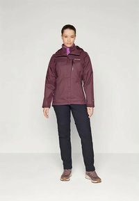 Woman standing wearing a purple hooded jacket, black pants, and purple sneakers against a plain light background.