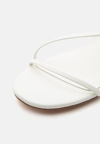 Μινιμαλιστικό λευκό σανδάλι με λεπτές λουράκια που διασταυρώνονται μπροστά και στρογγυλεμένη τετράγωνη μύτη σε ανοιχτό φόντο.