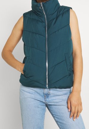 Veste matelassée sans manches de couleur teal foncé avec fermeture éclair frontale, col montant et poches, portée avec un jean bleu clair.