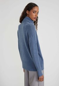 Donna con capelli ricci, che indossa un maglione a collo alto blu e pantaloni grigi, che guarda oltre la spalla contro uno sfondo neutro.