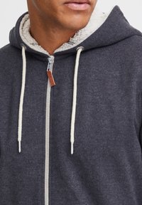 Sudadera con capucha y cremallera en tejido gris oscuro con forro de sherpa crema, que presenta cordones en contraste y detalle en etiqueta de cuero.