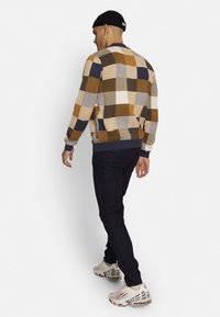 Strickpullover mit kariertem Muster in Braun, Creme, Marineblau und Senf; gerippte Bündchen und Saum; getragen mit schwarzen Jeans und Turnschuhen.