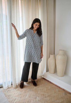 Frau in gemusterter Tunika und schwarzer Hose steht neben transparenten Vorhängen in der Nähe von zwei großen, beigen Keramikvasen auf einem gefliesten Boden.