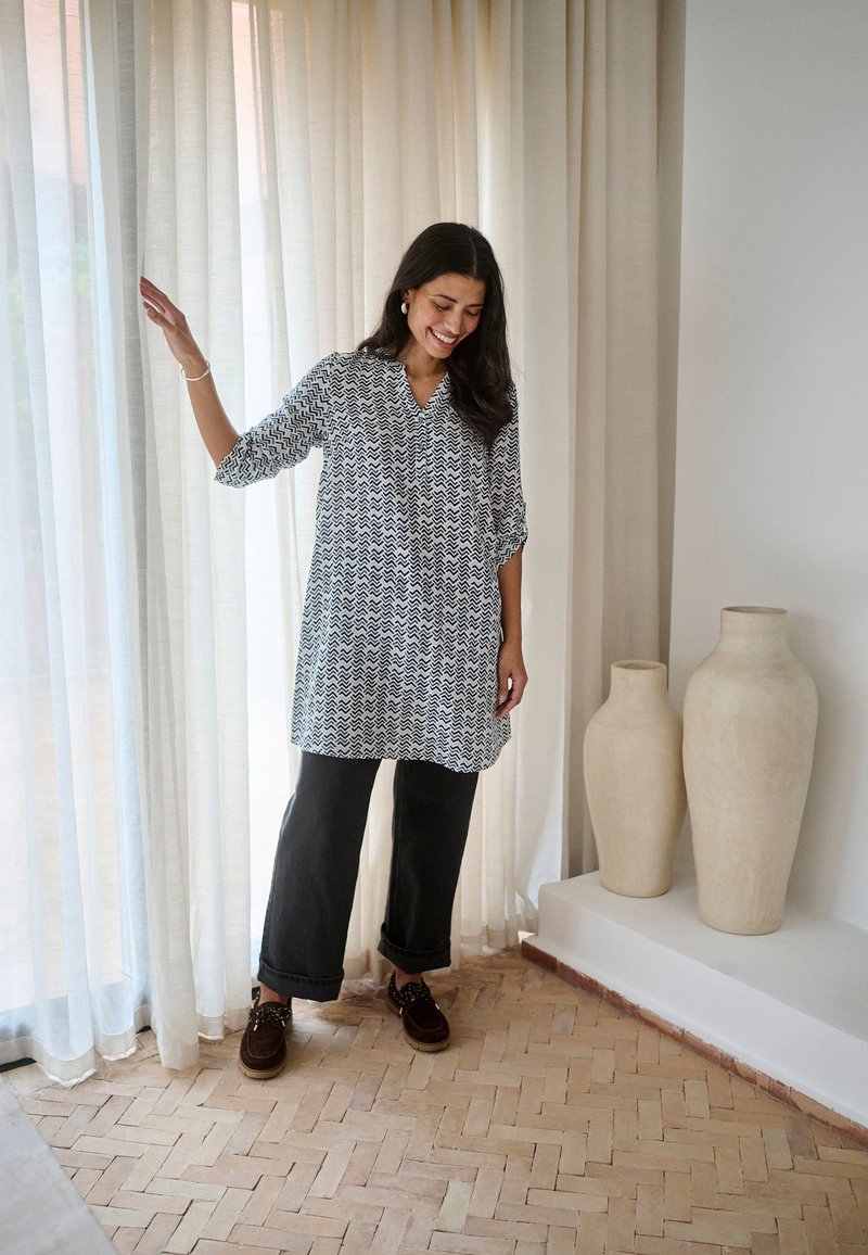 Naine mustriga tuunika ja mustade pükstega seisab läbinähtavate kardinate kõrval kahe suure beežika keraamilise vaasi juures plaatkattega põrandal.