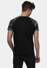 Svart t-skjorte med camo-ermer og rund hals. Kjemisk fiberblanding, tettsittende design, synlig tatovering på håndleddet. Minimalistisk stil.