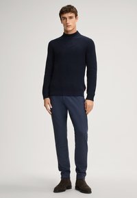 Marineblauer Ribbed Strickpullover mit hohem Kragen, kombiniert mit marineblauen tapered Hosen und dunkelbraunen Wildlederstiefeln. Einfaches, nahtloses Design.