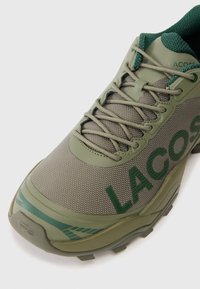 Olive green Lacoste sneaker z siateczkową cholewką, grubą podeszwą, wyściełanym kołnierzem, beżowymi sznurówkami i dużym tłoczonym logo z boku.