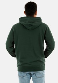 Sudadera verde con capucha y acabado texturizado, que cuenta con un bolsillo canguro y puños acanalados. Se lleva con unos jeans azul claro.