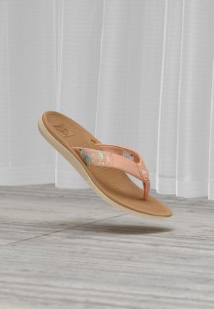 Sandale tannée type flip-flop avec des brides roses à motif floral flottant au-dessus d'un sol en bois gris et des rideaux blancs transparents en arrière-plan.
