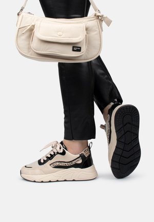 Person iført sorte bukser og beige sneakers med sorte accenter, bærende en beige skuldertaske med forlomme og knaplukning.