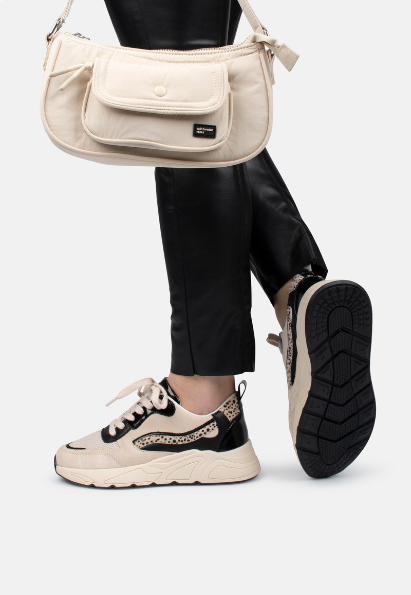 Personne portant un pantalon noir et des baskets beiges avec des accents noirs, portant un sac à bandoulière beige avec poche avant et fermeture à bouton.