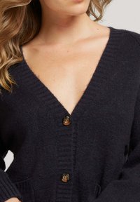Cardigan noir marine en maille douce, doté d'un décolleté en V profond, de poignets côtelés et de deux poches avant avec des boutons marron.