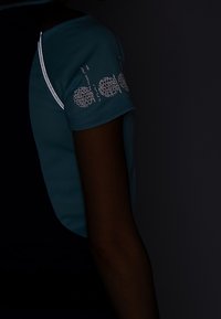 Short-sleeve athletic top in teal and navy with reflective accents on the shoulder. Features a patterned logo on the sleeve.