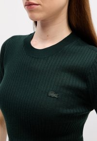 Zeleni pleteni kratki rukav top s okruglim izrezom. Sadrži mali, izvezeni tamnozeleni krokodil logo na lijevoj strani prsnog koša.