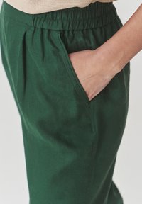 Pantalons en coton vert avec une ceinture élastique, présentant des poches latérales et une texture lisse. La jambe du pantalon comporte des plis subtils pour un détail supplémentaire.