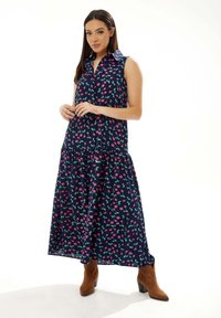 Navy sleeveless maxi dress with a floral print in pink and green, featuring a collar and tiered skirt. Model wearing brown ankle boots.