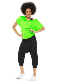 Neonově zelený oversized top v kombinaci s černými haremovými kalhotami, které mají elastické manžety. Modelka nosí bílé tenisky a má přirozeně strukturované vlasy.