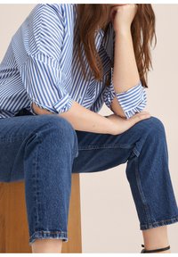 Chemise rayée bleu et blanc avec manches retroussées, associée à un jean court bleu foncé. La texture semble lisse avec une coupe décontractée.