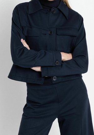 Personne portant une veste bleu marine foncée boutonnée avec des poches poitrine et un pantalon assorti, bras croisés sur la poitrine sur un fond clair.