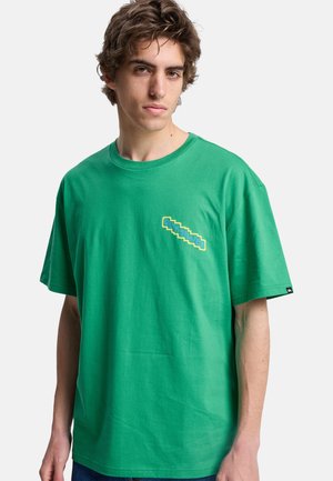 Młody mężczyzna z falowanymi brązowymi włosami, ubrany w zieloną koszulkę z krótkim rękawem z małym kolorowym logo na lewym ramieniu.