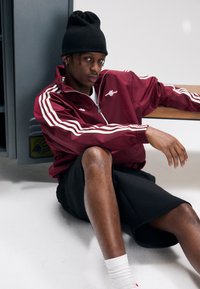 Casaco desportivo grená com riscas brancas e fecho de correr, combinado com calções pretos. Gorro preto. Modelo sentado contra um cacifo cinzento.