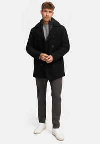 Schwarzer doppelt geknöpfter Peacoat mit großem Reverskragen, kombiniert mit einem grauen Strickpullover und grauen Hosen, abgerundet mit weißen Sneakers.