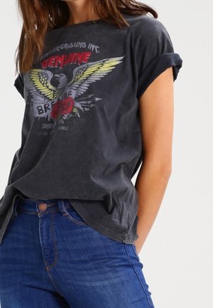 Vrouw draagt een zwart T-shirt met opgekrulde mouwen met een grafische opdruk van een adelaar met gele vleugels, rode rozen en blauwe jeans tegen een witte achtergrond.